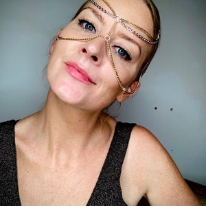 May include: A woman wearing a gold chain headpiece with a delicate design. The headpiece is made of multiple chains that are connected to form a geometric pattern. The woman is looking at the camera with a slight smile.