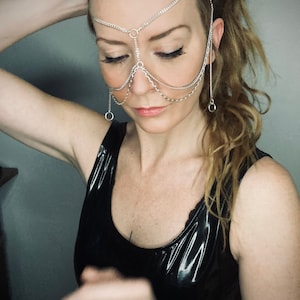 May include: A woman wearing a silver chain headpiece that covers her forehead, nose, and eyes. The chain is made of multiple strands and has small circular links. She is wearing a black tank top.