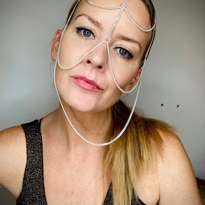 May include: A silver chain headpiece with multiple strands and circular accents. The headpiece is worn by a person with blonde hair and is positioned over the forehead and eyes.