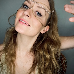May include: A woman with long, wavy, light brown hair is wearing a gold and silver chain headpiece. The chain headpiece covers her forehead and eyes. She is wearing a gold sparkly top.