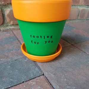 May include: A green and orange terracotta planter with the text "rooting for you" written in black.