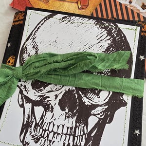 May include: A square card featuring a black and white graphic of a human skull, stitched with green thread onto a black background. A green fabric ribbon is tied horizontally across the center. A Halloween-themed napkin with a pumpkin is visible.