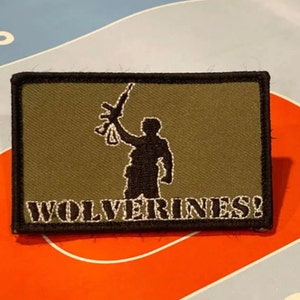 Pode incluir: Um remendo bordado verde e preto com a silhueta de uma pessoa segurando um rifle e o texto "WOLVERINES!"