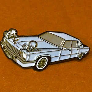 May include: A light blue car pin with a detailed design. The pin features a classic car with a white outline, and a brown background. The car has a detailed front grill, headlights, and wheels. The pin is likely made of metal.