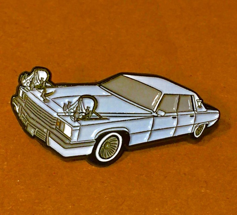 May include: A light blue car pin with a detailed design. The pin features a classic car with a white outline, and a brown background. The car has a detailed front grill, headlights, and wheels. The pin is likely made of metal.
