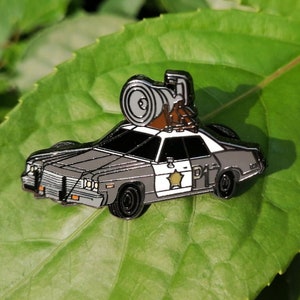May include: A black and white enamel pin of a police car with a large cannon on top. The car has a star and the number "D1" on the side.