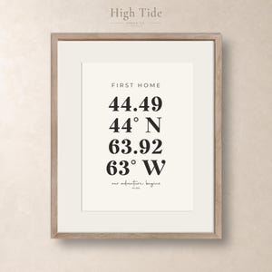 Peut inclure: Une affiche encadrée avec le texte FIRST HOME, les coordonnées 44.49 44° N 63.92 63° W et la phrase our adventure begins est. 2020 en noir sur fond crème, présentée dans un cadre en bois brun clair avec un passe-partout blanc.