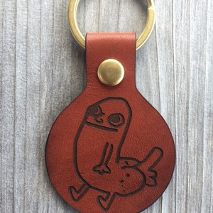 Pode incluir: Um chaveiro de couro marrom com um chaveiro de latão e uma etiqueta redonda de couro. A etiqueta tem um desenho de desenho animado de uma pessoa com uma cabeça grande e um corpo pequeno.
