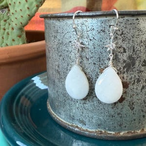May include: A pair of silver earrings with a starburst design and teardrop-shaped white stones. The earrings hang from silver hooks and are displayed against a textured, metallic background.