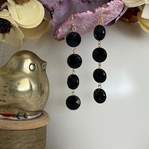 May include: A pair of dangle earrings featuring faceted black onyx beads suspended from gold-colored hooks. Each earring has four oval and round beads. The earrings are displayed against a white background with decorative elements.