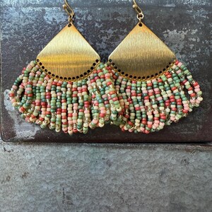 May include: A pair of handmade earrings with gold-colored, triangular tops and beaded fringe. The fringe features a mix of red, green, pink, and cream-colored beads, with gold-colored accents. The earrings are designed for pierced ears.