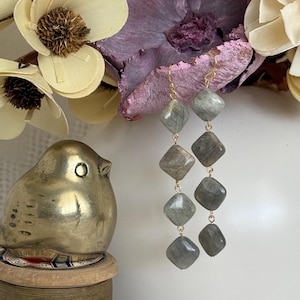 May include: Long dangle earrings featuring six gray, square-cut stones connected by gold-colored wire. The earrings hang in a vertical line. A gold-colored bird figurine and decorative flowers are in the background.