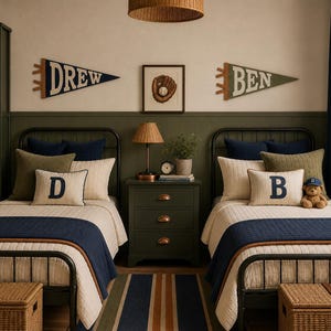 May include: A bedroom featuring two twin beds with black metal frames, cream-colored quilted bedding, and navy blue runners. Two felt pennants on the wall display the names DREW and BEN. A central green nightstand holds a lamp and a small plant.