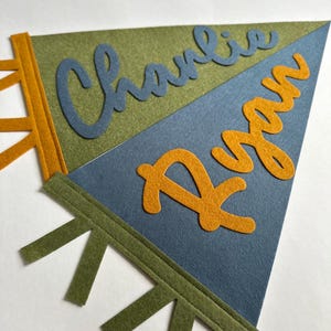 May include: Two triangular felt pennants featuring stitched details. The top olive green pennant displays the name Charlie in blue script, while the bottom blue pennant features the name Ryan in orange script. Both pennants have side tabs.