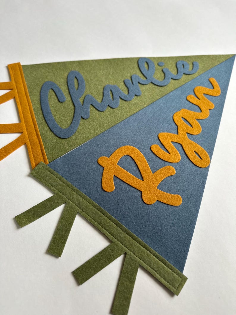 May include: Two triangular felt pennants featuring stitched details. The top olive green pennant displays the name Charlie in blue script, while the bottom blue pennant features the name Ryan in orange script. Both pennants have side tabs.