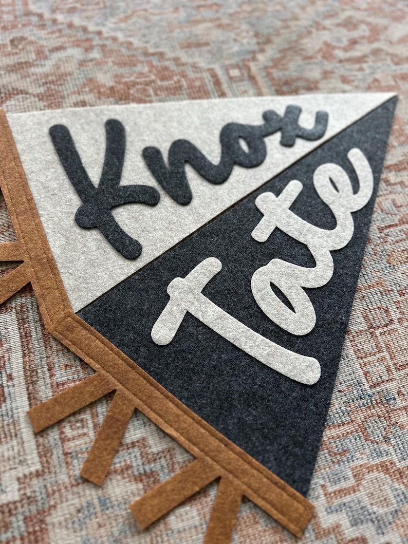 May include: A triangular felt pennant banner featuring a split design with a light gray top section and a dark charcoal bottom section. The text Knox is in dark charcoal felt, and the text Tate is in light gray felt. Brown felt fringe trim.