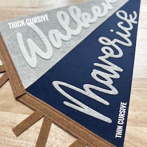 May include: A triangular felt pennant featuring a split design of light gray and dark navy blue. The word Walker is written in white thick cursive, and the word Maverick is written in white thin cursive. The pennant has brown felt tabs.