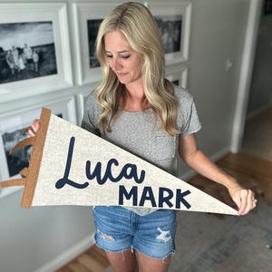 May include: A white felt pennant with the names "Luca" and "Mark" written in black cursive lettering. The pennant is being held by a person.