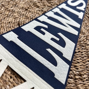 May include: A triangular navy blue felt pennant featuring the word LEWIS in large, bold, white capital letters. The pennant has a white border along the left side with decorative cutouts, showcasing a clean, high-contrast design.
