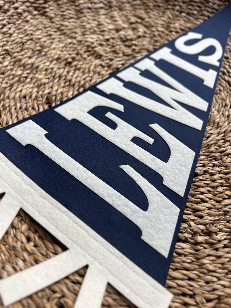 May include: A triangular navy blue felt pennant featuring the word LEWIS in large, bold, white capital letters. The pennant has a white border along the left side with decorative cutouts, showcasing a clean, high-contrast design.