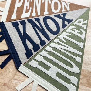 May include: Three triangular felt pennants featuring the names PENTON in white on a brown background, KNOX in dark blue on a light gray background, and HUNTER in white on an olive green background, each with matching felt ties at the base.