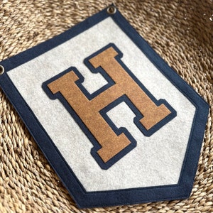 May include: A felt pennant banner with a large, brown felt letter "H" outlined in navy blue. The banner is a light gray color and has a navy blue border.