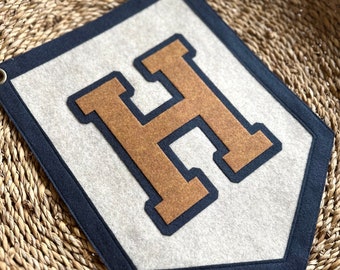 May include: A felt pennant banner with a large, brown felt letter "H" outlined in navy blue. The banner is a light gray color and has a navy blue border.