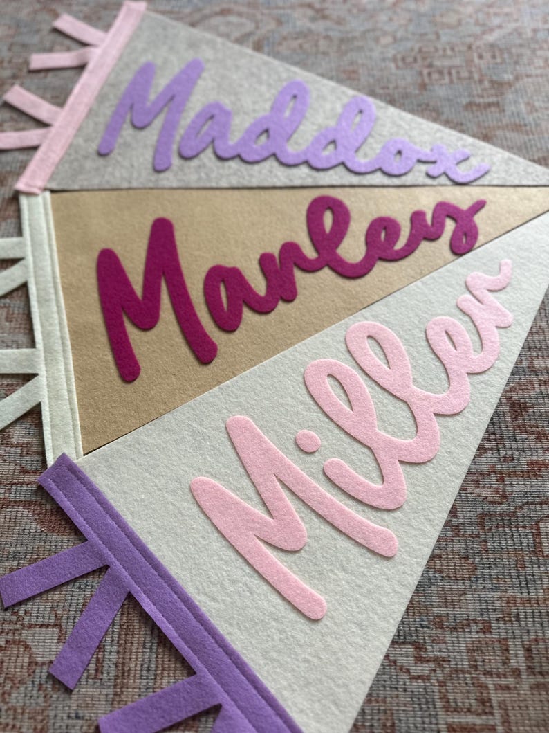 May include: Three triangular felt pennants with tabs on the left side. The top gray pennant features the purple text Maddox. The middle tan pennant features the dark pink text Manley. The bottom white pennant features the light pink text Miller.