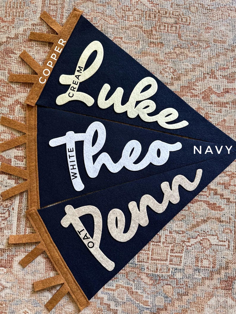 May include: Three triangular navy blue felt pennants with copper-colored fringe along the left side. Each pennant features a name in cursive felt lettering: Luke in cream, Theo in white, and Penn in oat. The text labels indicate the colors used.