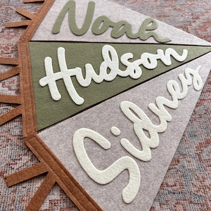 May include: A set of three triangular felt pennants arranged vertically, featuring the names Noah in olive green, Hudson in white, and Sidney in white. The pennants are light gray and olive green, with brown felt fringe trim along the left side.