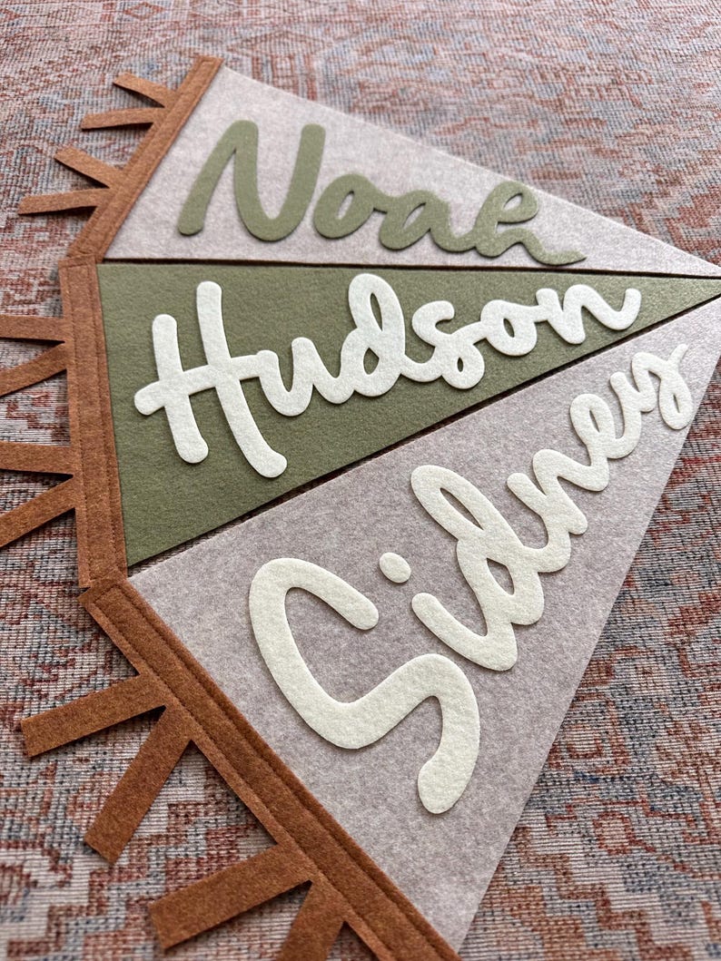 May include: A set of three triangular felt pennants arranged vertically, featuring the names Noah in olive green, Hudson in white, and Sidney in white. The pennants are light gray and olive green, with brown felt fringe trim along the left side.