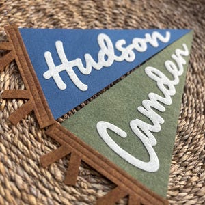 May include: Two triangular felt pennants featuring the names Hudson and Camden in white script lettering. The top pennant is blue and the bottom pennant is olive green. Both pennants have a brown felt trim with small, evenly spaced fringe tabs.