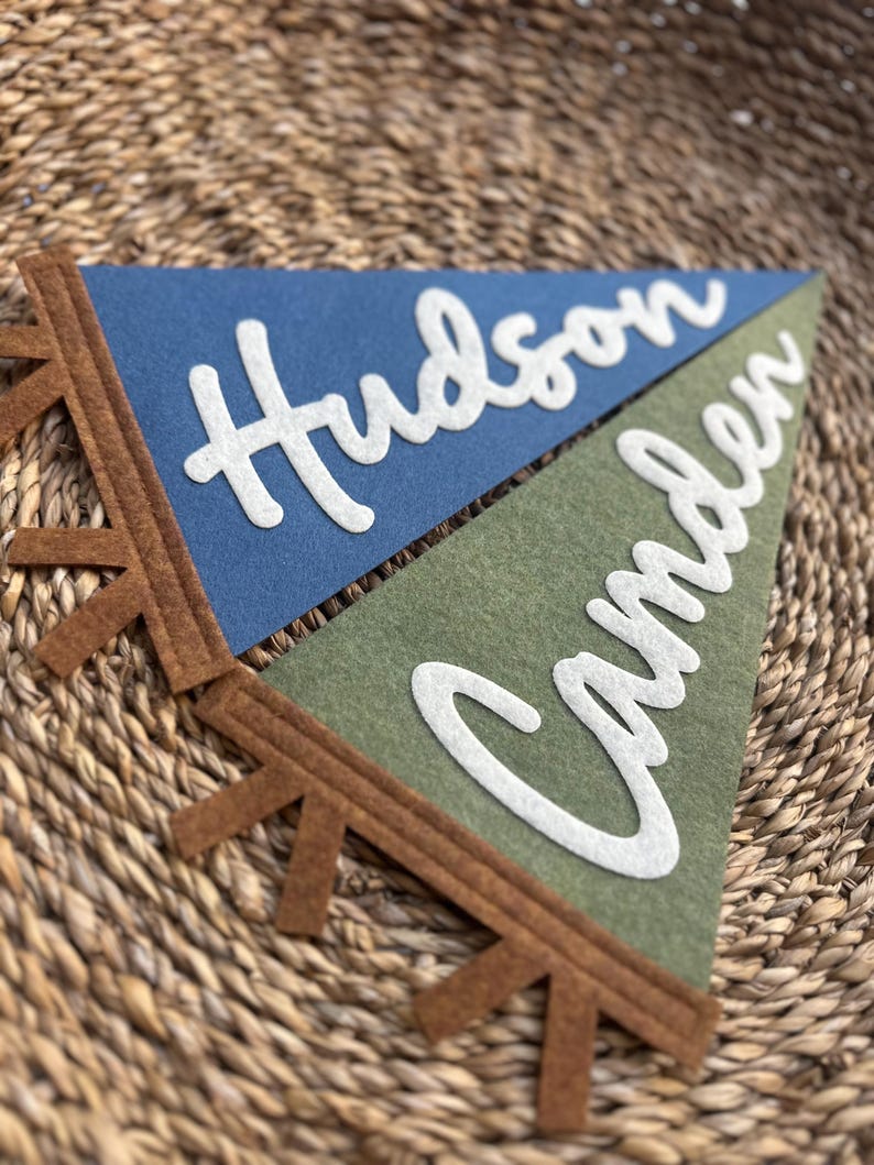 May include: Two triangular felt pennants featuring the names Hudson and Camden in white script lettering. The top pennant is blue and the bottom pennant is olive green. Both pennants have a brown felt trim with small, evenly spaced fringe tabs.