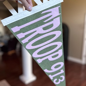 May include: A triangular olive green felt pennant featuring light pink felt lettering that reads TROOP 913. The pennant has a white felt header strip at the top with three vertical white felt tabs extending upward for hanging purposes.