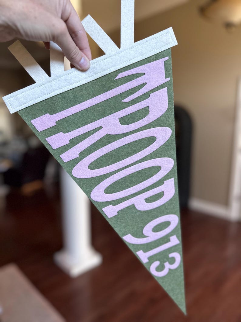 May include: A triangular olive green felt pennant featuring light pink felt lettering that reads TROOP 913. The pennant has a white felt header strip at the top with three vertical white felt tabs extending upward for hanging purposes.