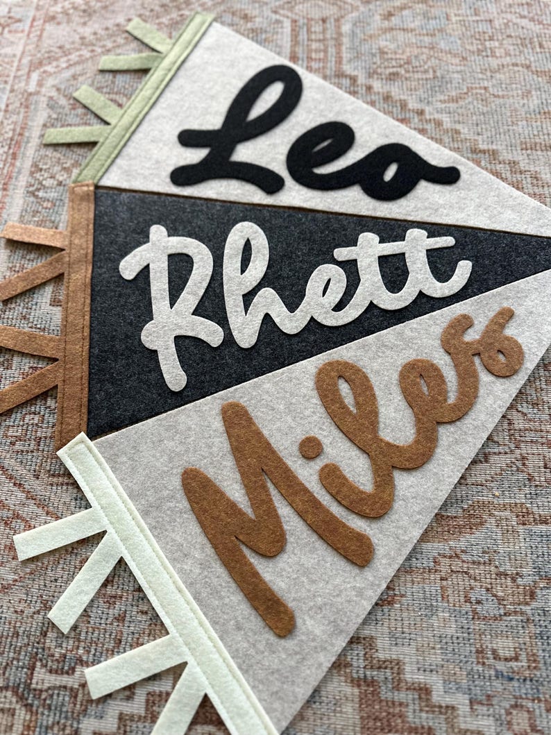 May include: Three triangular felt pennants layered together. The top pennant is light gray with the black felt name Leo. The middle pennant is dark charcoal gray with the white felt name Rhett. The bottom pennant is light gray with the brown felt name Miles.