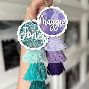Könnte beinhalten: Zwei weiße runde Ornamente mit gewellten Rändern und Quasten. Ein Ornament hat ein lila Marmormuster mit dem Namen "Maggie" in Weiß geschrieben. Das andere Ornament hat einen glitzernden, schillernden Hintergrund mit dem Namen "Jones" in Weiß geschrieben.