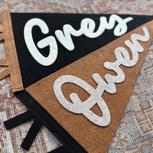 May include: Two triangular felt pennants featuring white cursive lettering. One pennant is black with the name Grey in white, and the other is brown with the name Gwen in white. Both pennants have contrasting colored felt tabs along the side.