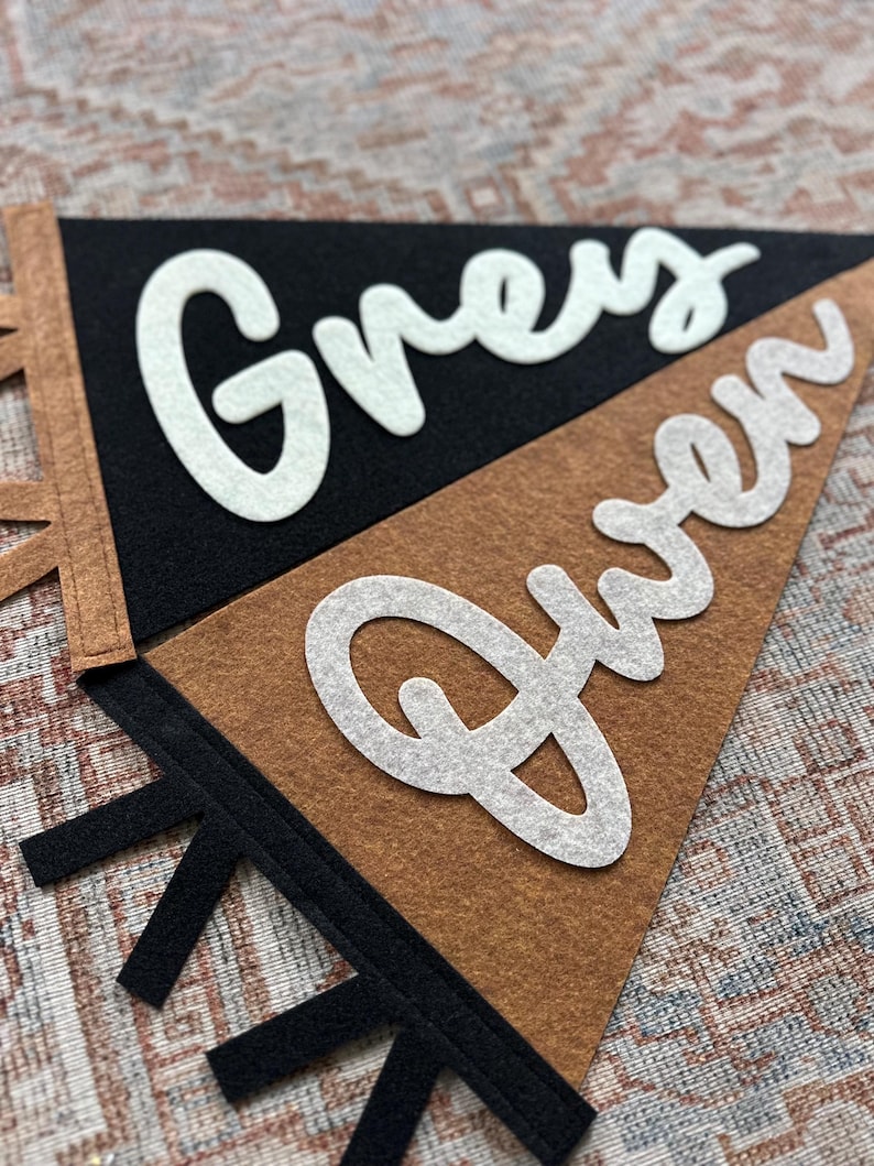 May include: Two triangular felt pennants featuring white cursive lettering. One pennant is black with the name Grey in white, and the other is brown with the name Gwen in white. Both pennants have contrasting colored felt tabs along the side.