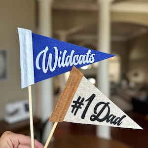 Può includere: Due bandierine in feltro su bastoncini di legno. Una è blu con "Wildcats" in corsivo bianco. L'altra è beige con "#1 Dad" in corsivo nero. Le bandiere sono triangolari e hanno un design classico a bandierina.
