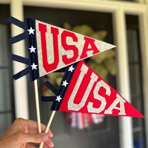 以下が含まれることがあります： 赤とグレーの「USA」の文字が入った、フェルト製のペナントフラッグ2枚。旗は赤い色にグレーの文字で、木のダボに取り付けられています。旗には、白い星が付いたネイビーブルーのアクセントがあります。