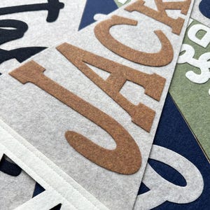 May include: A triangular felt pennant featuring the name JACK in large, brown, block-style letters. The pennant is light gray with a white stitched border at the top. Other overlapping felt pennants in navy blue, olive green, and white are visible.