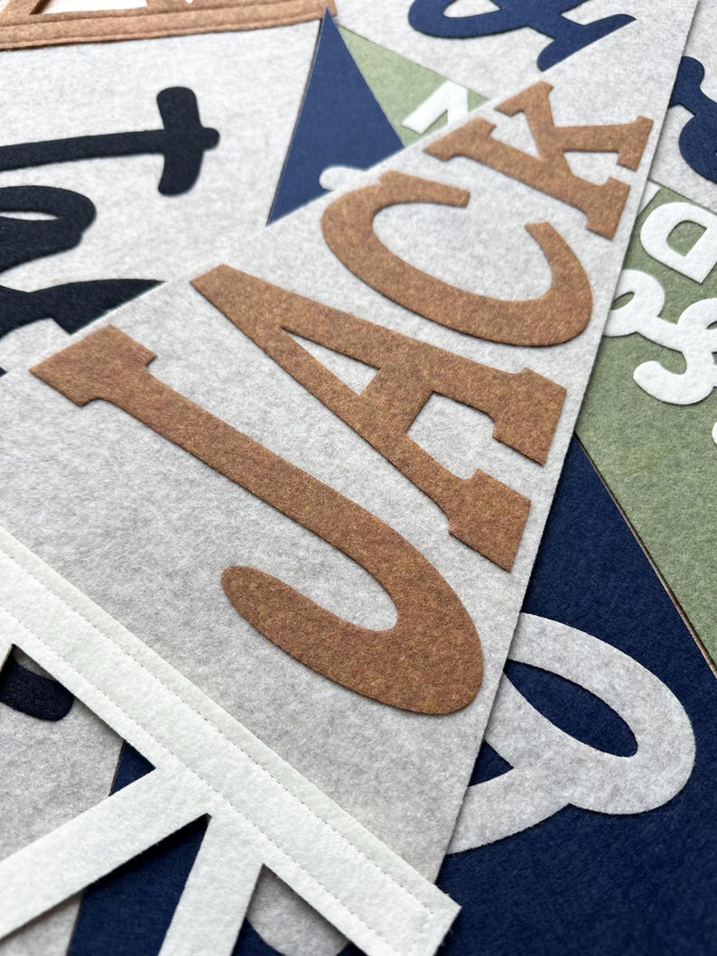 May include: A triangular felt pennant featuring the name JACK in large, brown, block-style letters. The pennant is light gray with a white stitched border at the top. Other overlapping felt pennants in navy blue, olive green, and white are visible.