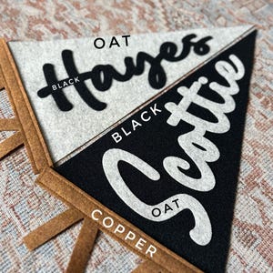 May include: Two triangular felt pennants with copper-colored tabs at the base. The top pennant is oat-colored with the black text "Hayes" in script. The bottom pennant is black with the white text "Scottie" in script. Labels indicate color options.