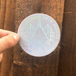 May include: A white, round sticker with a holographic finish. The sticker features the coat of arms of El Salvador, which includes a triangle with a mountain range, a volcano, and a sun. The text around the coat of arms reads "REPUBLICA DE EL SALVADOR EN LA AMERICA CENTRAL".