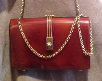 May include: A brown leather clutch purse with a gold chain strap and a gold clasp. The purse is closed and lying on a beige fabric surface.