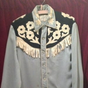 May include: A light blue long-sleeved shirt with a black and white embroidered design. The shirt has fringe trim and a button-up front.
