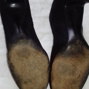 May include: A pair of black suede heeled shoes with worn leather soles. The shoes are placed on a white surface.