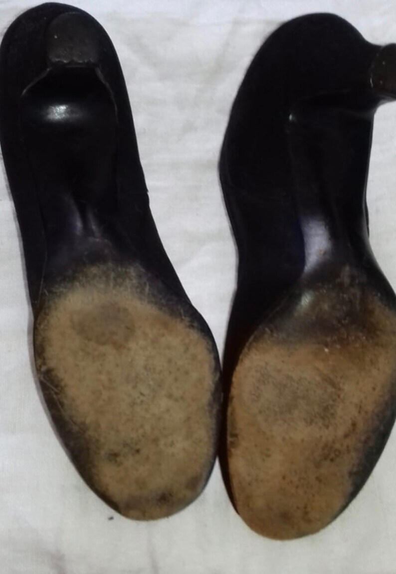May include: A pair of black suede heeled shoes with worn leather soles. The shoes are placed on a white surface.