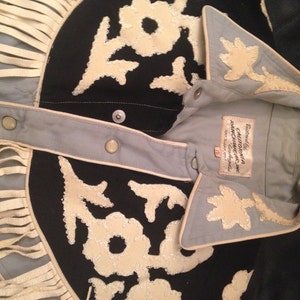 May include: A light blue denim shirt with a black and white embroidered design. The shirt has a white fringe trim and a white collar. The label reads "California Ranchwear Inc. Los Angeles."